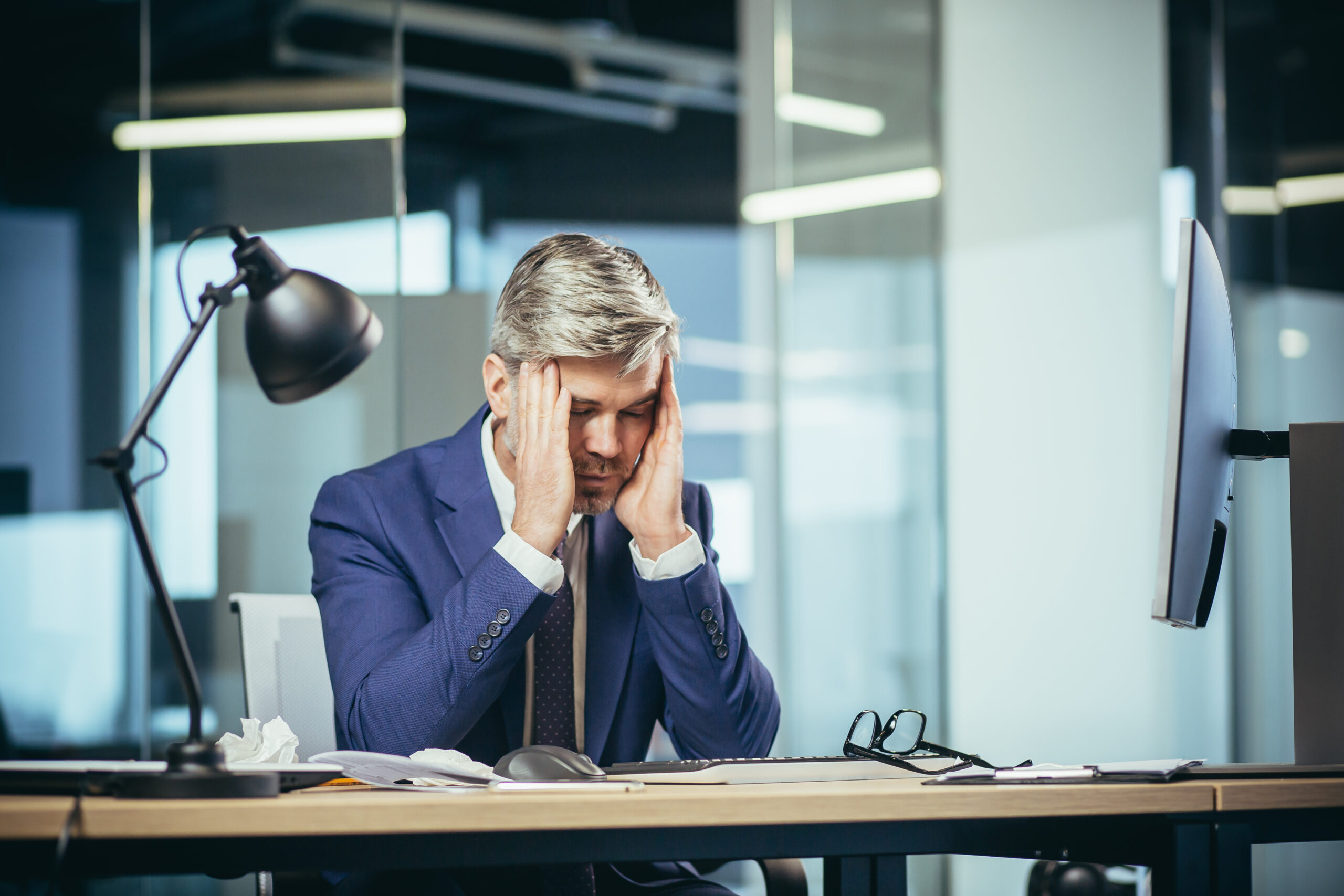 An experienced gray-haired businessman works at the computer until late, tired has a severe headache, stress and depression